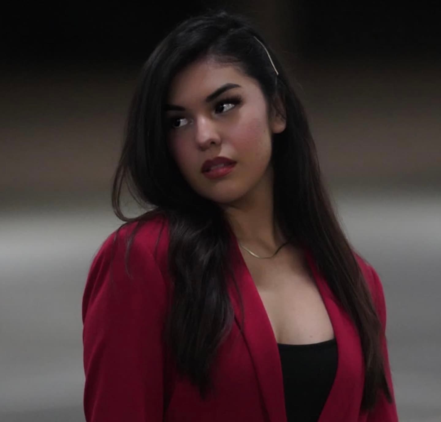 Young woman with long dark hair wearing a red blazer looking over her shoulder.